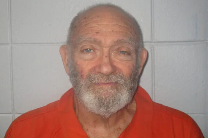 73-year-old man with a beard wearing an orange shirt, posing against a light gray tiled wall in a mugshot style.