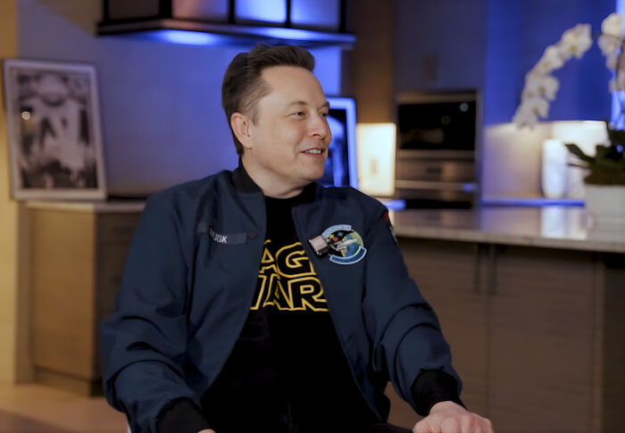 Elon Musk seated in a casual setting wearing a Space Wars shirt and dark jacket, engaged in conversation.