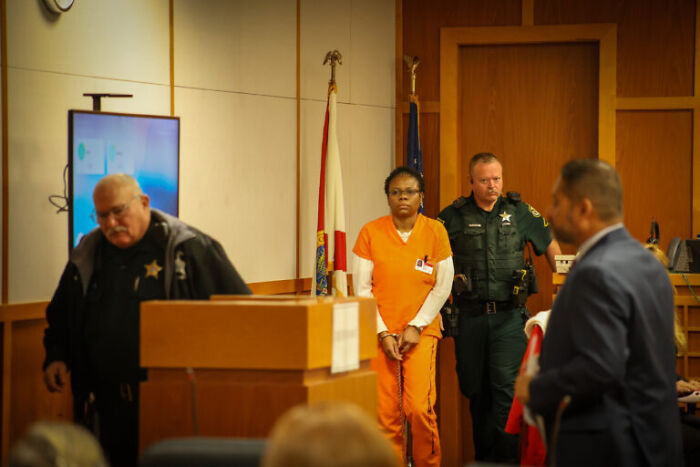 Woman in orange prison uniform escorted by sheriff deputies in courtroom during child a***e homicide case sentencing