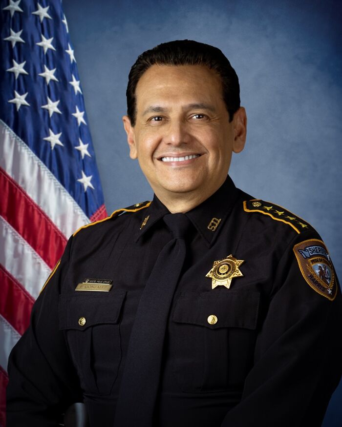 Texas sheriff smiling in uniform with badge and American flag in background representing law enforcement response.