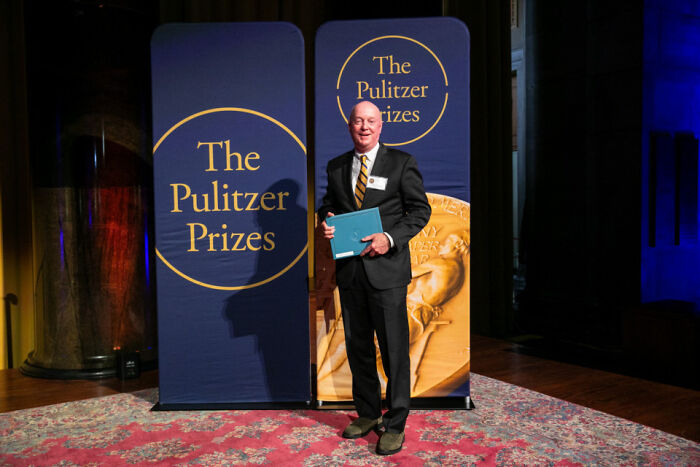 Man at Pulitzer Prizes backdrop holding folder, image referencing Trump and NYT Pulitzer photographer