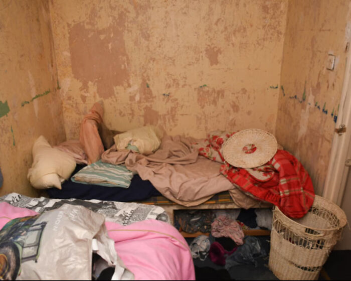 Cluttered small room with worn mattress, blankets, and laundry basket showing harsh conditions of vulnerable woman imprisoned.