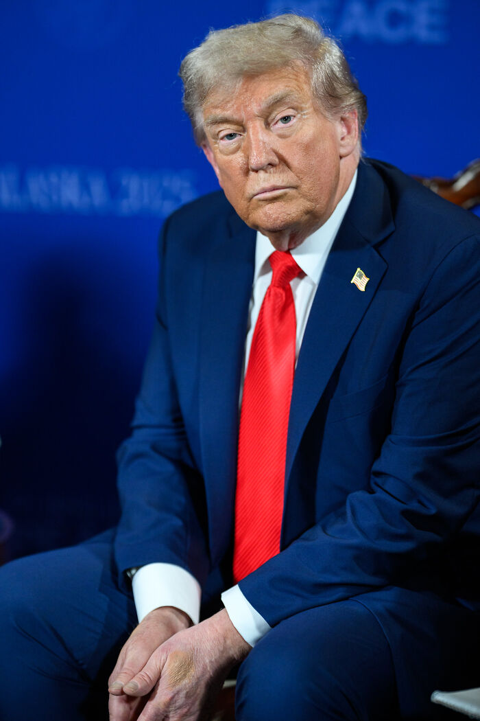 Donald Trump in a navy suit and red tie, sitting and looking serious while sending message about Renee Nicole Good shooting.