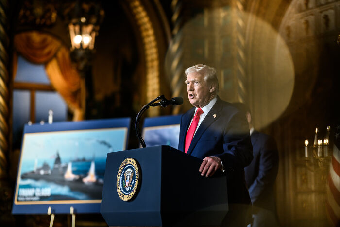 Former President Trump speaking at a podium with presidential seal, discussing Greenland residents wearing MAGA hats.