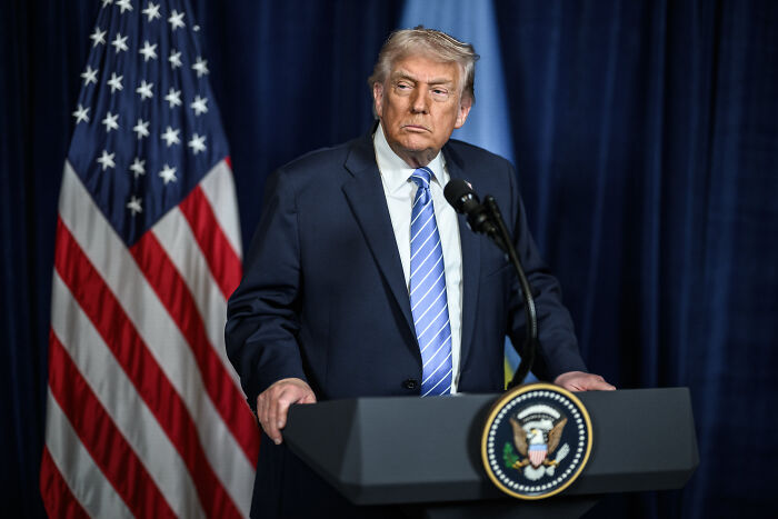 Former President Trump at a podium with US flag behind, discussing new price tag for dream military plans.