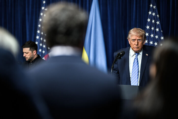 Preschool Teacher context: serious official at podium with US and other flags, blurred audience
