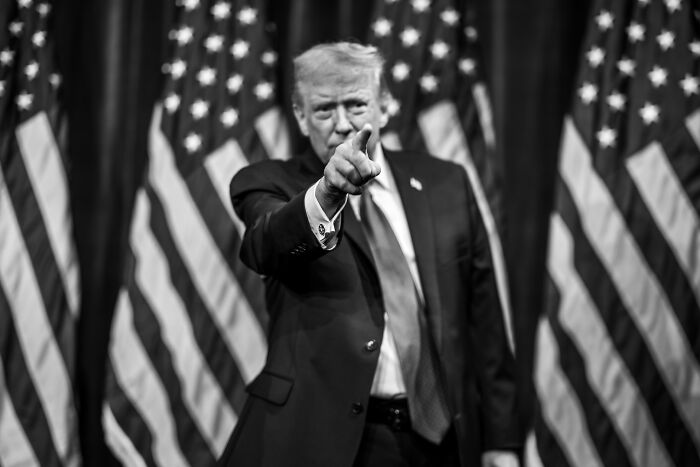 Person in a suit pointing forward with multiple American flags in the background during a political event involving White House war announcement.