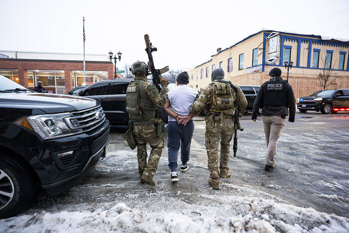 Heavily armed police escort a handcuffed individual outside a Texas ICE facility amid reports of children screaming inside.
