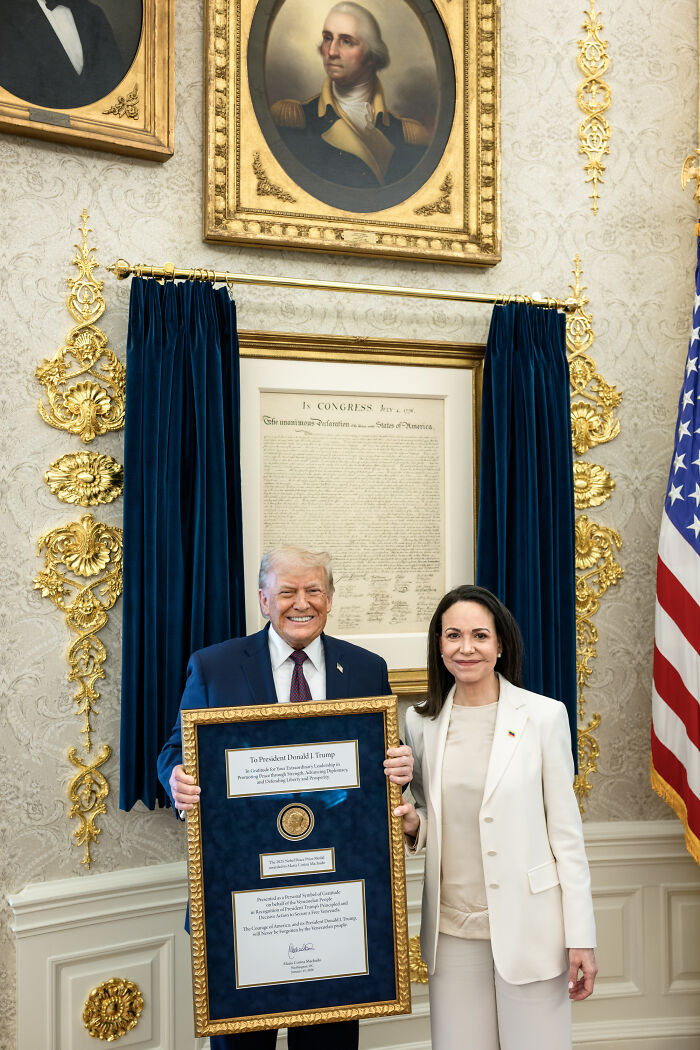 Donald Trump holding framed award with woman in elegant room featuring historical U.S. documents and American flag.