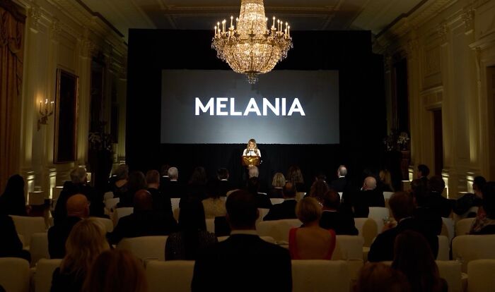 Melania Trump at a movie event presenting her film titled Melania to a seated audience in an elegant room.