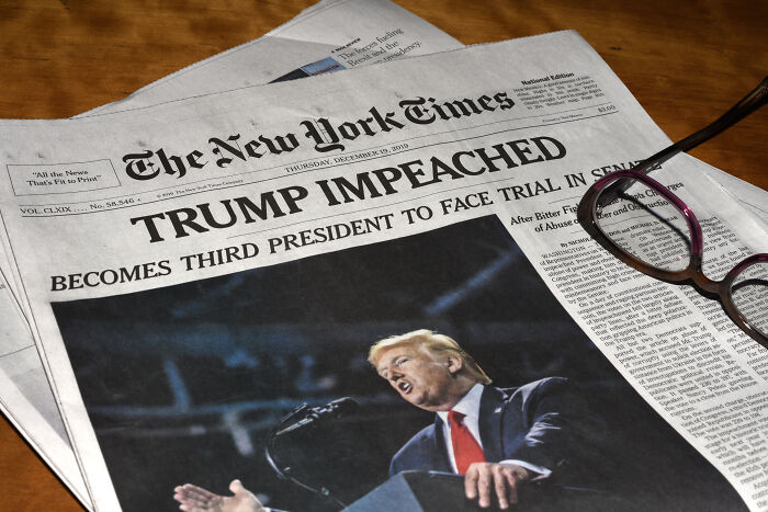 Newspaper front page with large IMPEACHED headline, photo of politician speaking, glasses beside paper