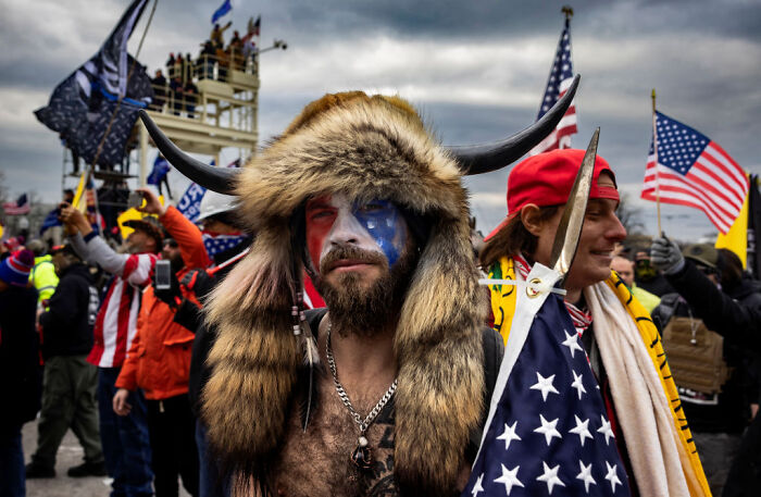 Man in horned fur headdress and face paint holding US flag amid political rally crowd