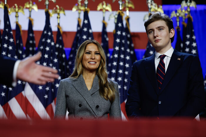 Melania Trump and Barron Trump standing in front of multiple American flags at a formal event.