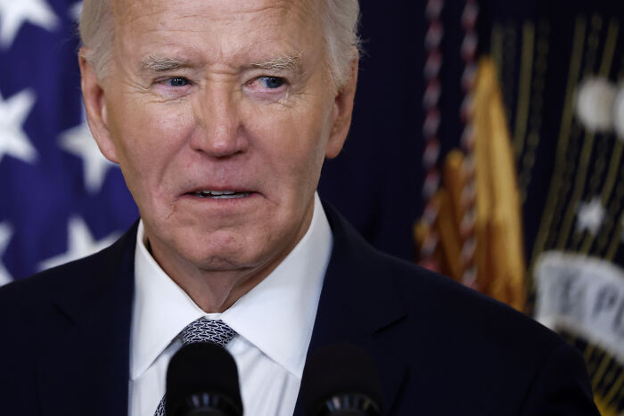 Close-up of a serious man speaking at a podium with American flags in the background, showing a tense expression.