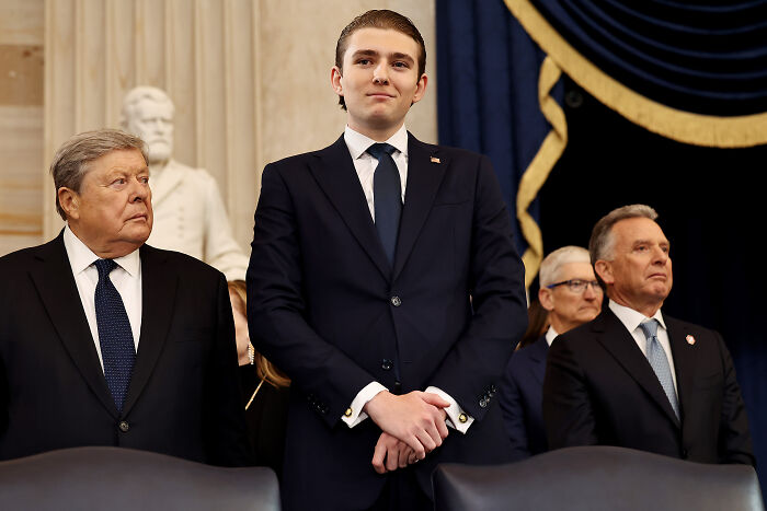 Barron Trump in a navy suit standing among men in formal attire at a ceremonial or official event indoors.