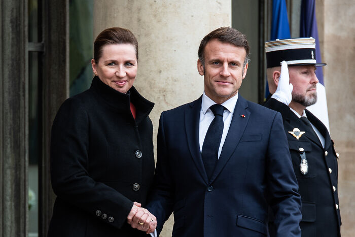 Two people shaking hands in formal attire with a uniformed guard in the background during a public event.