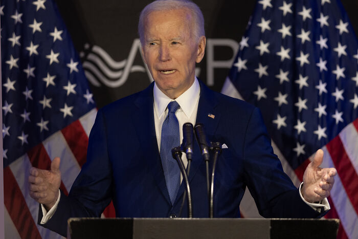Joe Biden speaking at a podium with American flags in the background during a speech about Trump raves and impersonation.