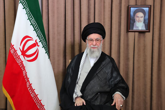 Senior Iranian cleric sitting next to Iranian flag with framed portrait behind, related to U.S. locked and loaded warning to Iran.