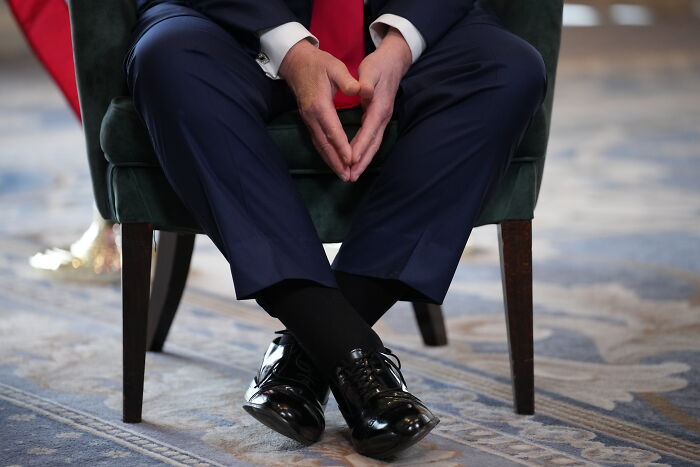 Man sitting cross-legged in polished black shoes and dark compression socks, wearing a navy suit and red tie, health-related.