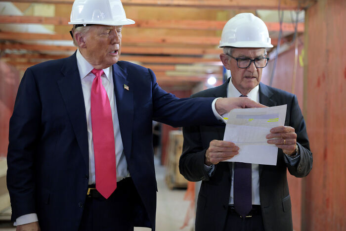 Donald Trump and Fed Chair Jerome Powell wearing white hard hats reviewing documents at a construction site.