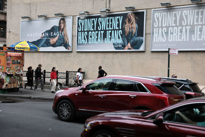 Billboards featuring Sydney Sweeney promoting jeans as pedestrians walk by on a busy city street with cars in traffic.