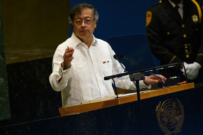 Man in white shirt speaking at United Nations podium with security officer standing behind, discussing geopolitical tensions.