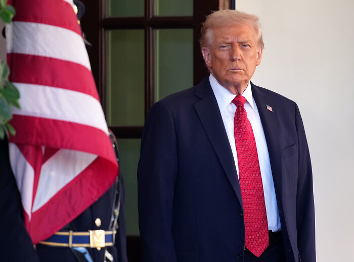 Man in navy suit and red tie standing beside U.S. flag, NYT Pulitzer mentioned