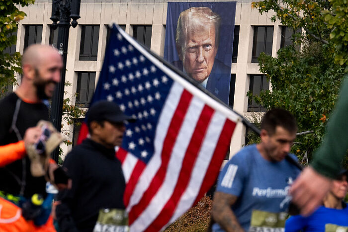 People holding an American flag during a public event with a large portrait of Donald Trump in the background.