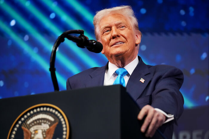 Donald Trump speaking at a podium with the presidential seal, representing Americans and American identity after his second year.