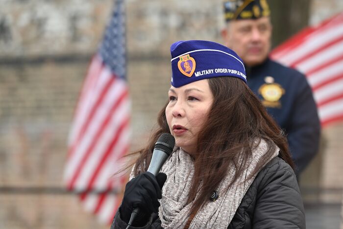 Woman in military hat speaking into microphone at outdoor event with American flags, related to combat veteran controversy.