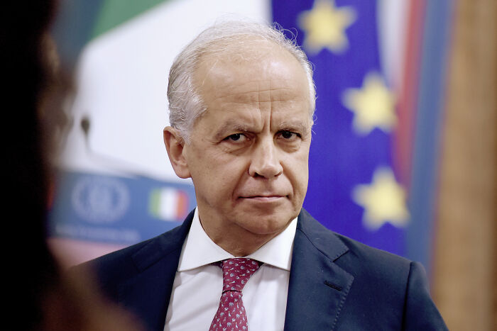 Older man in suit and red tie standing indoors with blurred flags in the background, related to ICE deployment at Olympics.