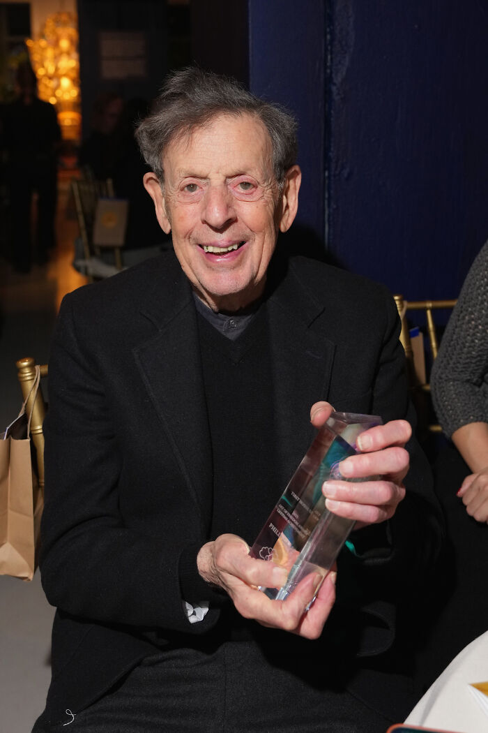 Philip Glass smiling and holding an award in a black suit, related to pulling new symphony from Kennedy Center.