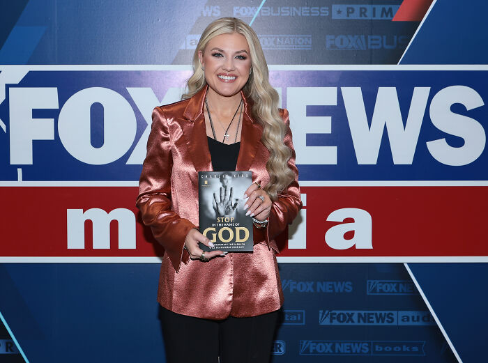 Erika Kirk smiling in front of Fox News media backdrop holding a book amid viral audio controversy.