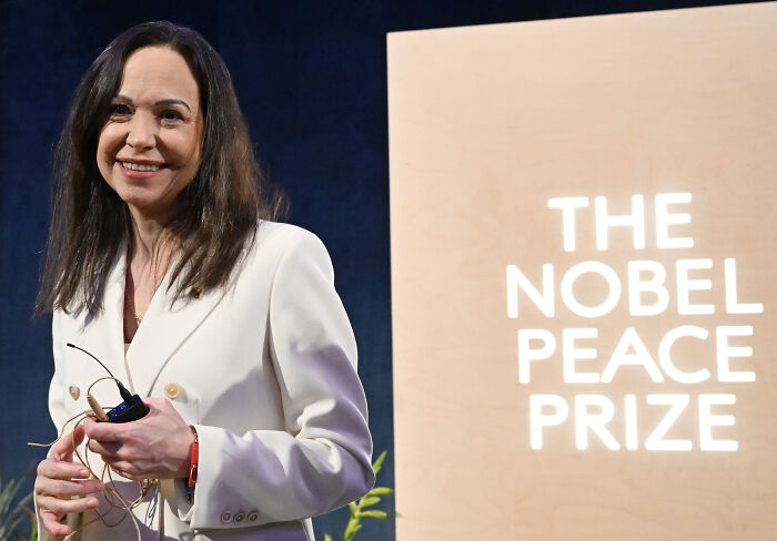 Woman in a white blazer holding a microphone near a sign reading The Nobel Peace Prize, related to Trump and Machado peace prize offer.