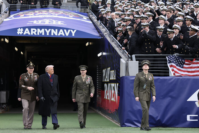 Donald Trump walking with military officers near a stadium tunnel with soldiers watching a football game event.