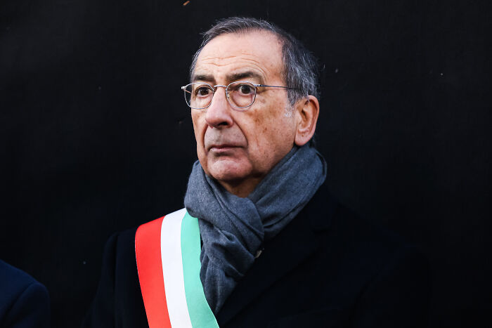 Man wearing glasses, dark coat, and scarf with an Italian flag sash, looking serious against a black background.