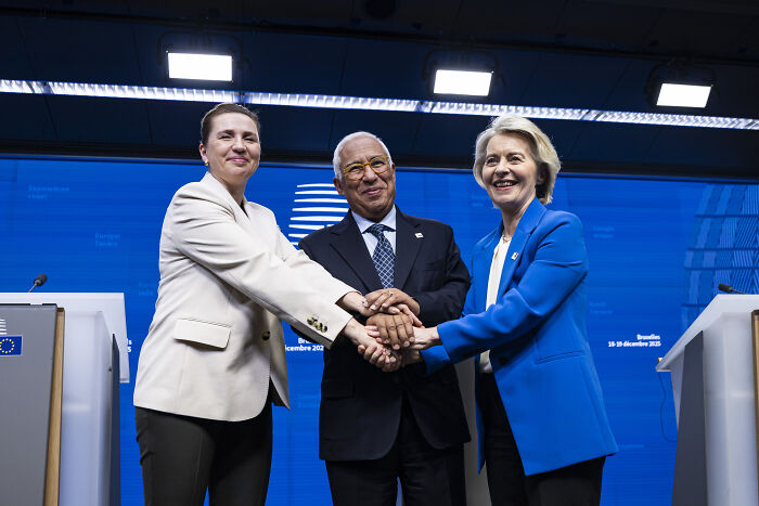 Three leaders shaking hands at a press conference with Greenland residents wearing MAGA hats in the background.