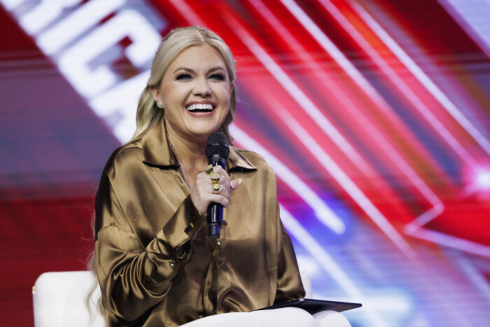 Erika Kirk speaking into microphone, smiling during an event with colorful blurred background. Audio of Erika Kirk viral.