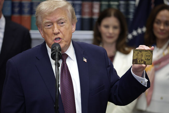 Donald Trump wearing a suit and tie, speaking at a microphone, holding a gold card with his image and signature.