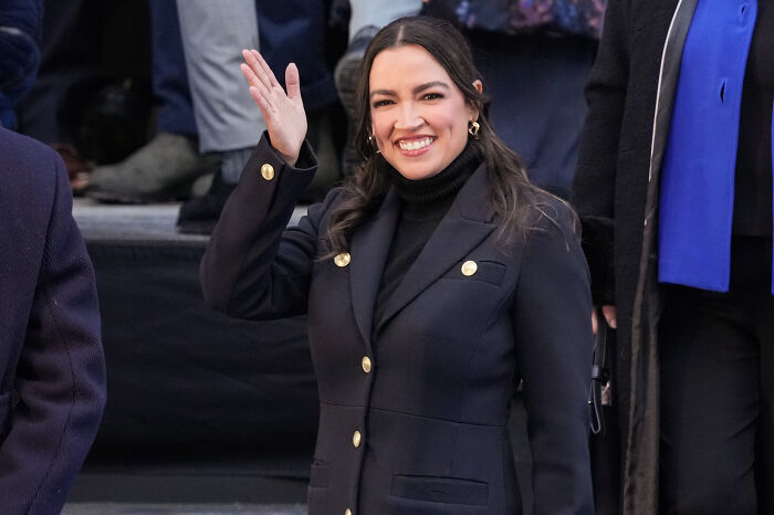 Alexandria Ocasio-Cortez smiling and waving, dressed in a black coat, during a public political event on cold weather.