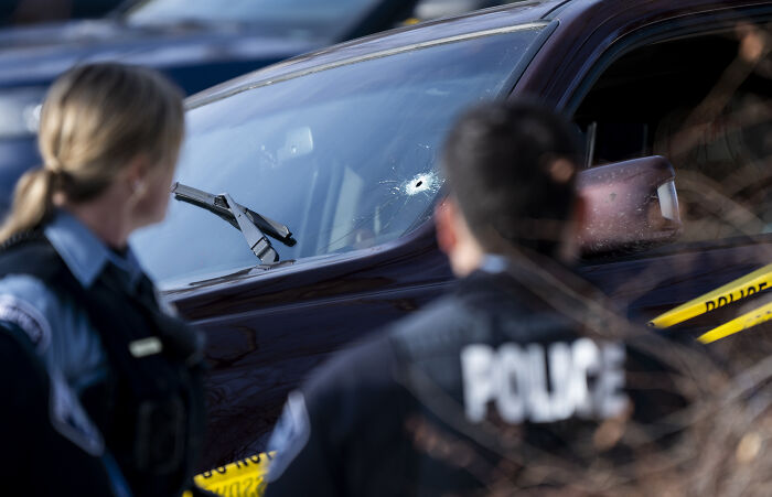 Police officers examine a vehicle with a bullet hole in the windshield at a crime scene related to ICE incident.