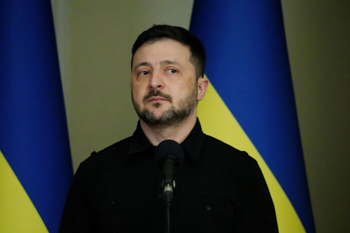 Volodymyr Zelenskyy speaking seriously at a podium with Ukrainian flags in the background during a press event.