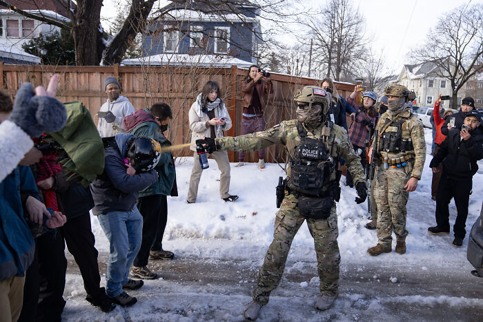 Police in tactical gear using pepper spray to disperse protesters near snowy Minneapolis streets, relating to Renee Good&rsquo;s training disruption.
