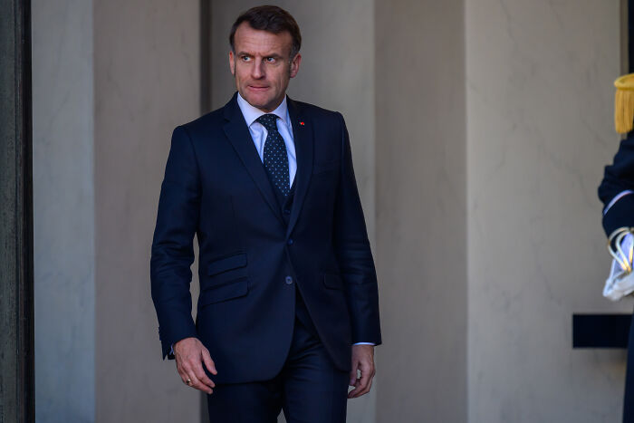 Man in a dark suit, representing Macron, near an entrance, related to wine tariffs and political tensions.