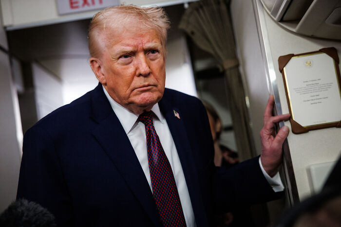 Man in a dark suit and red tie standing inside an aircraft, with a serious expression and holding a wall plaque.