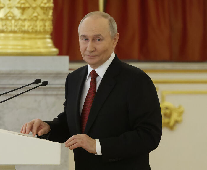Vladimir Putin in a formal suit speaking at a podium during an official event focused on geopolitical issues.