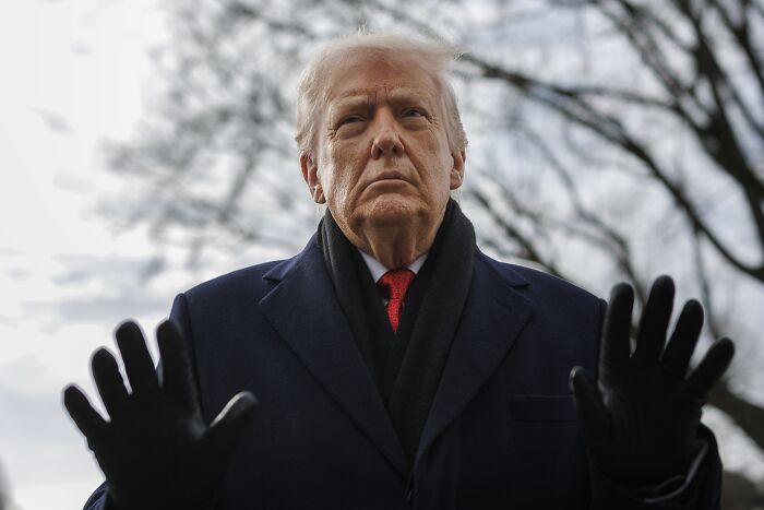 Former President Trump outdoors at Davos, wearing a dark coat and gloves, with a serious expression and raised hands.