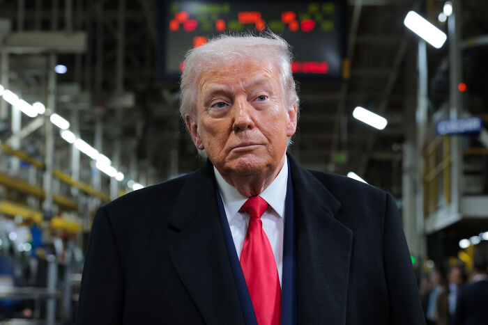 Donald Trump in a dark industrial setting, wearing a black coat and red tie, related to MAGA support and campaign efforts.