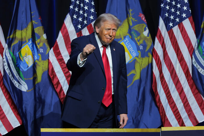 Donald Trump on stage, wearing a suit with red tie, gesturing during a speech with American flags in the background.
