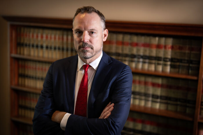 Man in navy suit and red tie standing with arms crossed in front of law books, related to Minnesota prosecutors investigation.